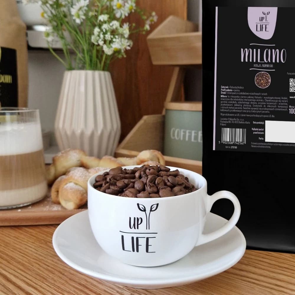 Filiżanka z logo UpLife wypełniona po brzeg ziarnami kawy Milano, obok opakowanie 1 kg i rogale na desce